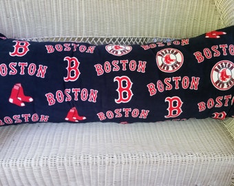 May include: A long, rectangular pillow with a navy blue fabric featuring a repeating pattern of red "Boston" text, red "B" logos, and red and white baseball socks. The pillow is sitting on a white wicker chair.