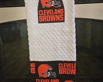 May include: A white textured kitchen towel featuring an embroidered Cleveland Browns helmet logo and team name patch at the top, with a black fabric band at the bottom printed with repeating orange Cleveland Browns helmet logos and text.