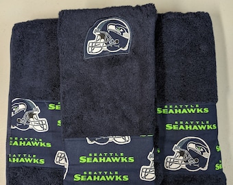 Puede incluir: Tres toallas de color azul marino con un logotipo de los Seattle Seahawks en verde y blanco. Las toallas tienen un patrón repetido del logotipo en la parte inferior.
