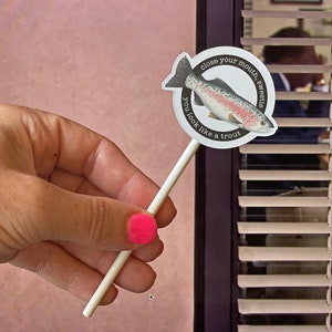 May include: A round white sticker on a stick features a watercolor illustration of a trout. The sticker has the text "close your mouth, sweetie, you look like a trout" in a circular pattern around the fish.