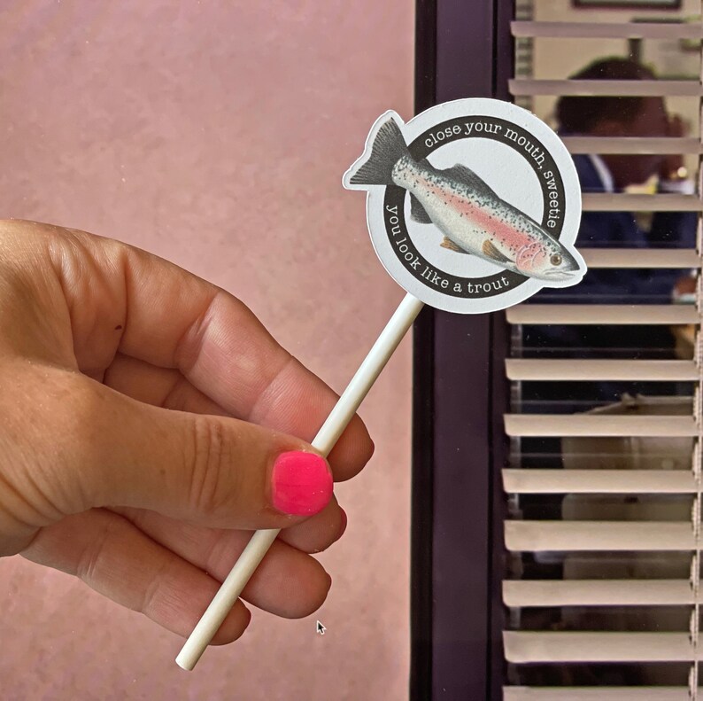 May include: A round white sticker on a stick features a watercolor illustration of a trout. The sticker has the text "close your mouth, sweetie, you look like a trout" in a circular pattern around the fish.