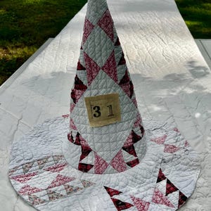 May include: A conical hat constructed from quilted fabric featuring a white base with red and dark pink geometric star patterns. A rectangular beige fabric patch is stitched to the front of the cone, displaying the black printed number 31.