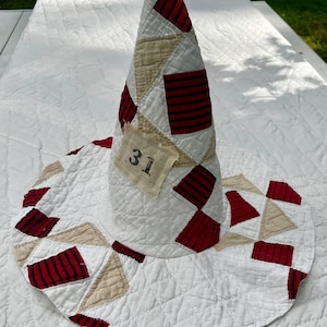 May include: A quilted cone-shaped hat featuring a patchwork design of white, beige, and dark red striped fabric squares. A small rectangular fabric patch with the black printed number 31 is stitched onto the side of the conical crown.