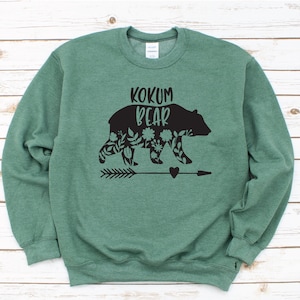 Può includere: Una felpa verde con un disegno di orso floreale nero e il testo "Kokum Bear" stampato sul davanti.