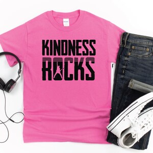 May include: Pink t-shirt with black text that reads "Kindness Rocks" with a silhouette of a person playing guitar.