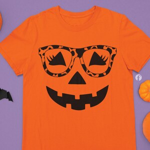 May include: Orange t-shirt with a black pumpkin face design wearing leopard print sunglasses. The pumpkin has a wide smile and a small nose.
