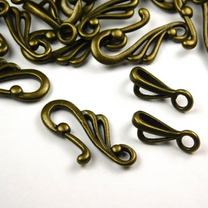 May include: Antique bronze metal clasps with ornate designs. The clasps have a hook shape with a decorative swirl pattern.