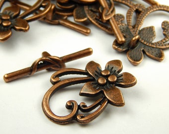 May include: Copper-toned metal toggle clasp with a floral design. The clasp features a decorative swirl and a flower with three smaller flowers in the center.