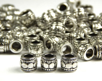 May include: Silver-colored metal beads with a cylindrical shape and decorative patterns. The beads have a series of raised dots and circular designs. A collection of these beads are scattered on a white surface.