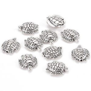 May include: A collection of silver-colored turtle-shaped beads. Each bead features a detailed shell pattern and small head and flipper details. The beads are scattered on a white surface, ideal for jewelry making or crafting.