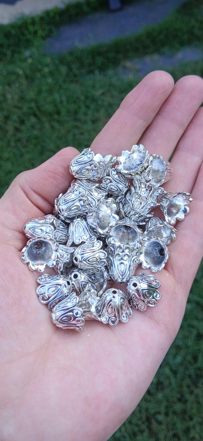 Op de afbeelding: Een verzameling kleine, zilverkleurige metalen kraalkapjes in de vorm van klokvormige bloemen. Elk stuk is voorzien van ingewikkelde, in reli&euml;f gemaakte krulpatronen en een centraal gat aan de bovenkant, ontworpen voor het maken van sieraden en knutselprojecten.