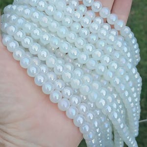 May include: Strands of iridescent white beads. The beads are round and have a pearlescent sheen, reflecting a rainbow of colors. The beads are strung together, ready for jewelry making or other crafts.