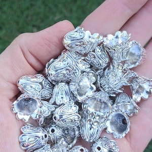 Op de afbeelding: Een verzameling zilverkleurige metalen kraalkappen met een gedetailleerd, reli&euml;f bloem- of bloembladpatroon. Elk stuk is gevormd als een kleine, open bel of bloemknop met een centraal gat aan de bovenkant om ze aan een draad te rijgen.