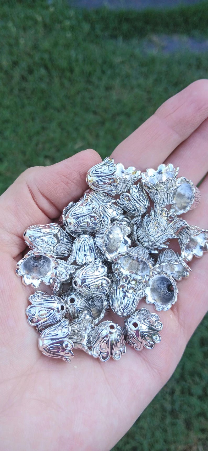 Op de afbeelding: Een verzameling zilverkleurige metalen kraalkappen met een gedetailleerd, reli&euml;f bloem- of bloembladpatroon. Elk stuk is gevormd als een kleine, open bel of bloemknop met een centraal gat aan de bovenkant om ze aan een draad te rijgen.