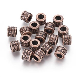 May include: A collection of bronze-colored metal beads with intricate embossed designs. The beads are cylindrical with a central hole and feature swirling and geometric patterns. The beads are scattered on a white surface, showcasing their detailed craftsmanship.