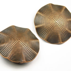 May include: Two copper-colored, spiral-patterned decorative objects. Each object has a unique, irregular shape with concentric circles radiating from the center. The objects are set against a white background, highlighting their metallic sheen.