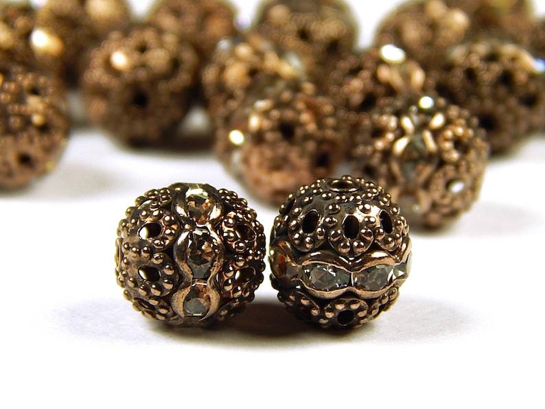 May include: Two ornate gold-toned metal beads with clear rhinestone accents. The beads have a decorative, filigree design.