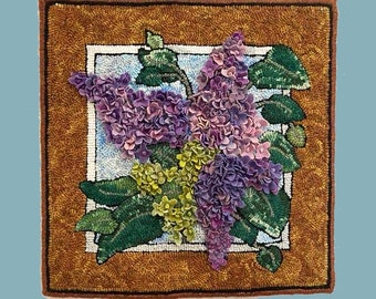 May include: A close-up of a needlepoint rug depicting a bouquet of purple and pink lilac flowers with green leaves. The rug is framed with a brown border and a white inner frame. The text "BeachNutStudio ©" is in the lower left corner.