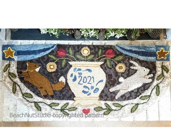 May include: A hand-stitched rug with a brown, white, and blue vase with the year 2021 on it. The vase is surrounded by flowers and leaves. A white rabbit and a brown fox are on either side of the vase. The rug has a blue and white banner at the top with a star on each end. The rug is on a white brick surface. BeachNutStudio copyrighted pattern.