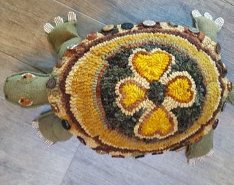 May include: A green and brown stuffed turtle with a patterned shell. The shell is made of yarn and has a design of three yellow hearts in the center. The turtle has button eyes and a button on its neck.
