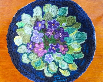 May include: A round, blue wool rug with a floral design. The rug features a bouquet of purple, pink, and white flowers with green leaves. The rug is hand-hooked and has a textured surface.