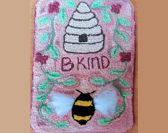May include: A rectangular rug with a pink background features a beehive, a bee, and the words "B KIND." The bee has yellow and black stripes and white wings. Green vines with red flowers frame the top and sides of the rug.