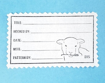 May include: A white rectangular label with a scalloped edge, featuring a line drawing of a sheep holding a crochet hook. The label includes fields for "TITLE:", "HOOKED BY:", "DATE:", "NOTE:", and "PATTERN BY:" on a light blue background.
