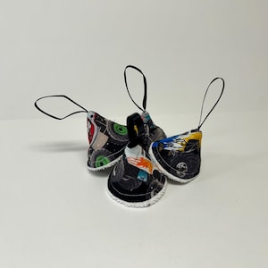 May include: Four fabric ornaments with black loops for hanging. The ornaments are shaped like bells and feature a colorful monster truck tire pattern. The edges are trimmed with white stitching.