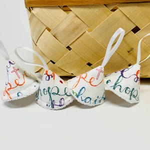 May include: Three white fabric ornaments with the words "hope" and "faith" in colorful script, each with a small cross design. The ornaments have white ribbon loops for hanging and are in front of a woven basket.