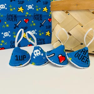 Puede incluir: Cuatro adornos de tela azul con ribete blanco y lazos de cinta blanca. Cada adorno presenta gráficos pixelados de calavera, corazón, espada y estrellas, con el texto "1UP" y "GAME OVER". Un tejido azul con gráficos similares está en el fondo.