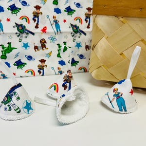 May include: Three cone-shaped fabric ornaments with white terry cloth interiors and white hanging loops. The exterior fabric features a repeating pattern of cartoon characters, rainbows, stars, and rockets in blue, green, red, and yellow colors.