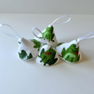 May include: Five white fabric ornaments with green frog designs and white ribbon loops. The ornaments are cone-shaped and have a white background with green frogs printed on them.