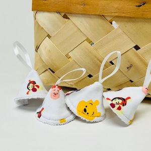 May include: Four white fabric bell ornaments with cartoon character designs, each with a white ribbon loop. The characters include Winnie the Pooh, Piglet, and Tigger. A woven basket is in the background.