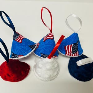 May include: A collection of six fabric ornaments with hanging ribbons. Three are cone-shaped with a blue background and American flag print. Three are circular with textured fabric in solid red, white, and blue, each featuring a matching ribbon loop.