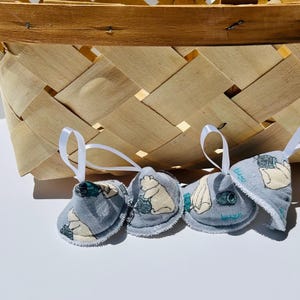 May include: Three small, grey fabric ornaments with white ribbon loops hang in front of a woven basket. Each ornament features a different illustration in cream and teal. The basket is made of light brown woven wood.