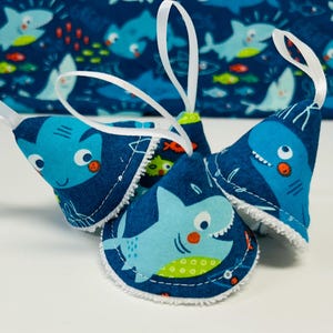 May include: Four blue shark-themed ornaments with white ribbon hangers. Each ornament features a different shark design with white trim. The background shows a blue fabric with a repeating shark and fish pattern.