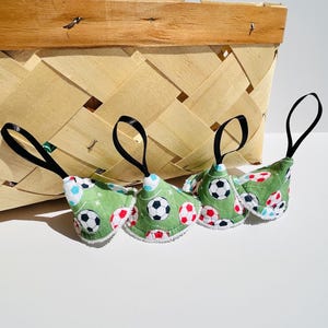 May include: Four green fabric ornaments with soccer ball patterns and black loops hang in front of a woven basket. The ornaments have white trim and are decorated with black, red, and blue soccer balls.