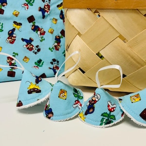 Puede incluir: Artículos de tela azul claro con temática de Super Mario Bros. La tela presenta a Mario, champiñones y otros elementos del juego. Se adjuntan lazos de cinta blanca. Una cesta tejida está en el fondo.