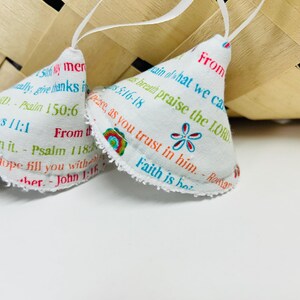 May include: White fabric ornaments with colorful text and floral designs. The ornaments have a bell shape and are trimmed with a white, frayed edge. Each ornament has a white ribbon for hanging. A woven basket is in the background.