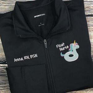 May include: Black zip-up jacket with the text "Anna, RN, BSN" embroidered on the left chest. The right chest features an embroidered design of a unicorn float with the words "Float Nurse". The jacket has a high collar and two front pockets.