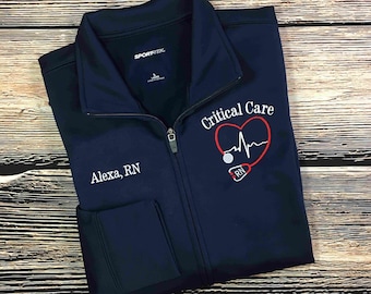 May include: A navy blue zippered jacket with the text "Critical Care" embroidered on the right side of the chest. A red heart with a stethoscope and an EKG line is embroidered inside the text. The text "Alexa, RN" is embroidered on the left side of the chest.