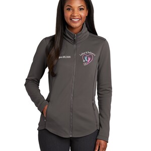 May include: A charcoal gray women's zip-up jacket with a small embroidered logo on the left chest. The logo features a pink heart with two blue footprints inside and the text "Labor & Delivery". The text "[Name] RN, BSN" is also embroidered on the jacket.