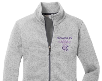 May include: A gray fleece zip-up jacket with a pocket on the left side. The jacket has a small embroidered design on the right chest that includes a caduceus symbol and the text "Sharonda, RN".