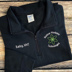May include: A navy blue quarter-zip sweatshirt with the text "Bailey, NMT" embroidered on the left chest. The right chest features "Nuclear Medicine Technologist" with a green atom graphic. The sweatshirt is on a wooden surface.