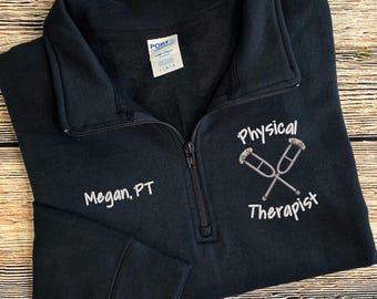 May include: A navy blue quarter-zip sweatshirt with the text "Megan, PT" embroidered on the left chest. The right chest features the words "Physical Therapist" with crossed crutches. The sweatshirt is made of a soft material.