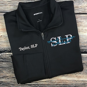 May include: Black Sport-Tek zip-up jacket with the text "Taylor, SLP" embroidered on the left chest. The right chest features the words "SLP Speech Language Pathologist" in white and teal. The jacket has a full zipper and two front pockets.