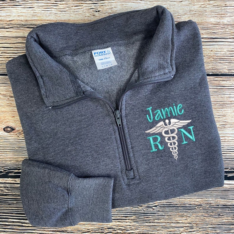 May include: A dark gray fleece pullover with a zipper. The pullover has a white embroidered medical symbol with the text "Jarmie RN" in teal.