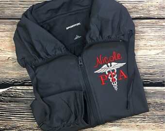 May include: A dark gray zippered jacket with a hood. The jacket has a white embroidered medical symbol and the text "Nicole PTA" in red.