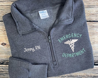 Op de afbeelding: Een donkergrijze trui met rits met de tekst "Emergency Department" geborduurd in groen en wit. De tekst "Jenny, RN" is in wit onder de rits geborduurd.
