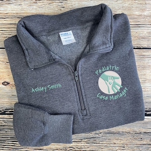 May include: A dark gray zippered sweatshirt with the text "Pediatric Case Manager" and a green hand logo embroidered on the right side. The text "Ashley Smith" is embroidered on the left side.